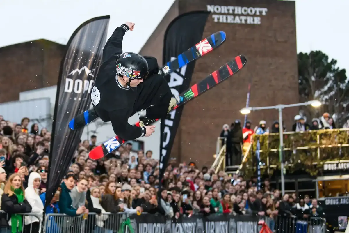 Skier grabbing their skis mid-air above the Chill Hill crowd, Northcott Theatre behind