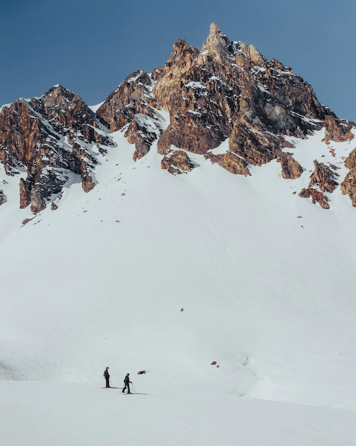 Two skiers dwarfed by a jagged peak above a wide snowfield