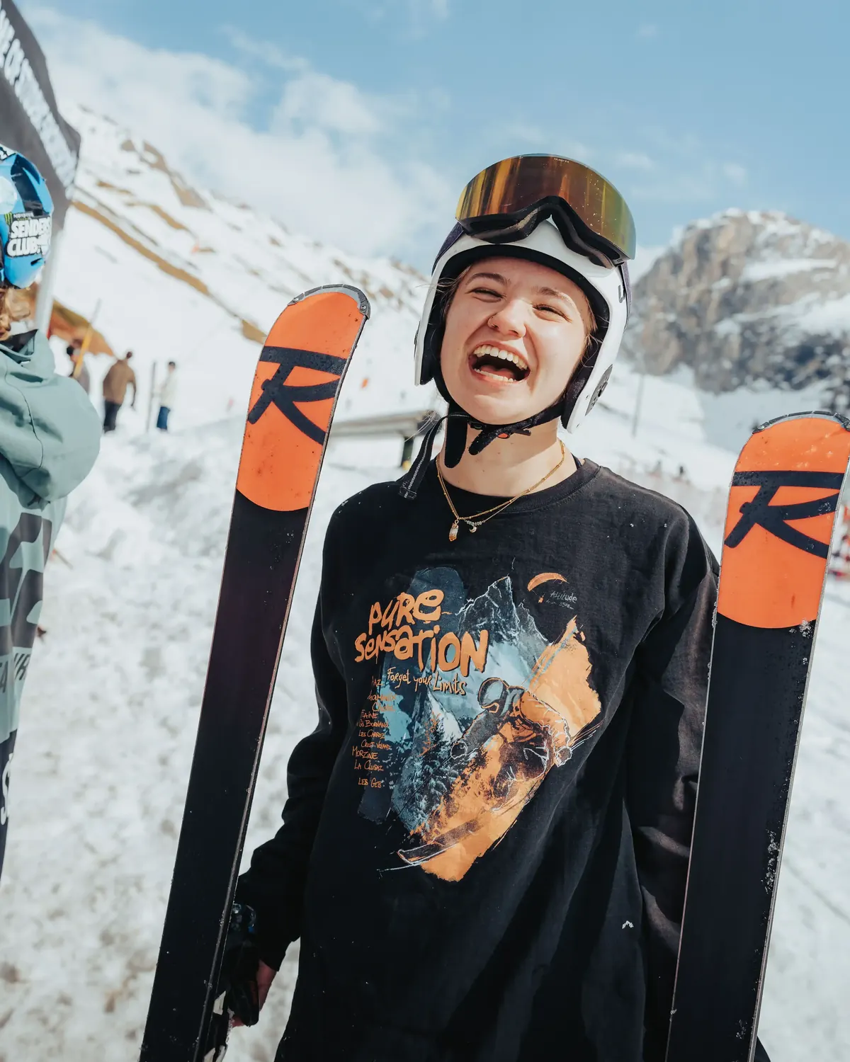 Member laughing while carrying their skis, mountains behind